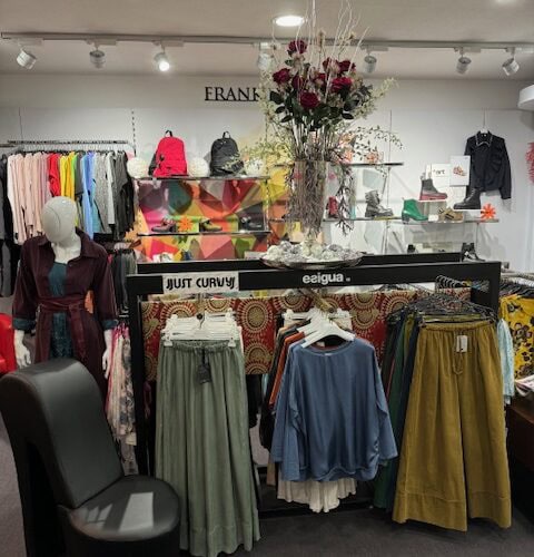Ein Modegeschäft mit bunten Kleidungsstücken, einer Blumenarrangement und einem leeren schwarzen Stuhl.